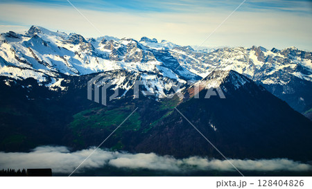 Majestic snow-capped mountains rise above serene green valleys under a clear blue sky in the Swiss Alps during midday 128404826
