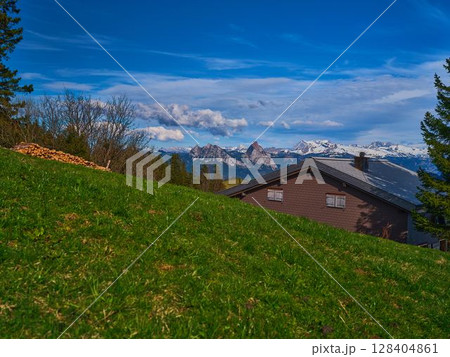 Majestic mountain landscape viewed from a grassy hill with a cozy cabin under a clear blue sky 128404861