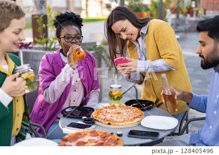 Friends enjoying pizza and drinks together at an outdoor restaurant, capturing moments with a phone. 128405496