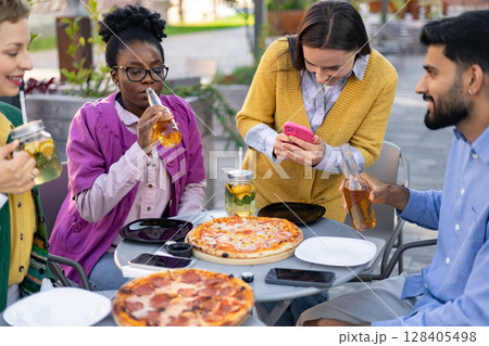 Friends enjoying pizza and drinks at an outdoor cafe, capturing moments with a phone and having a good time together. Friends enjoying pizza and drinks at an outdoor cafe, capturing moments with a phone and having a good time together. 128405498
