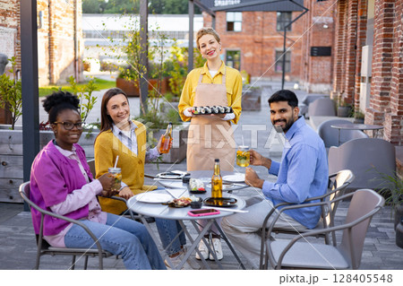 Diverse group of friends enjoying sushi and drinks at an outdoor restaurant, served by a friendly waitress. Diverse group of friends enjoying sushi and drinks at an outdoor restaurant, served by a friendly waitress. 128405548