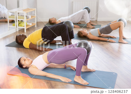 Multinational group of people lie on mats, hold Pilates ball between their knees and perform abdominal exercises during group exercise in fitness studio Multinational group of people lie on mats, hold Pilates ball between their knees and perform abdominal exercises during group exercise in fitness studio 128406237