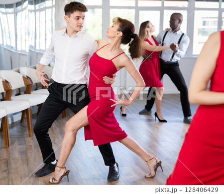 Beautiful young woman in red dress at dance salon with her partner dancing tango 128406429