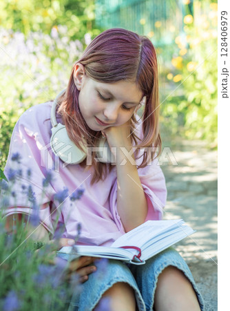 Young girl reading a book in a garden while wearing headphones Young girl reading a book in a garden while wearing headphones 128406979