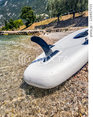Stand-up paddleboard on pebble beach beside turquoise Lake Garda waters. High quality photo 128407555