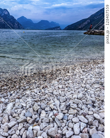 Rocky shore of Lake Garda with clear water and mountains under cloudy sky. High quality photo 128407559