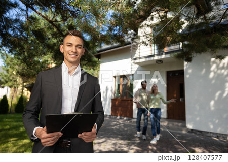 Real estate agent with clipboard outside modern house with couple 128407577