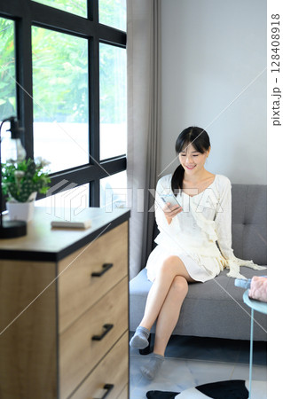 Asian woman in a white dress sitting on a sofa and using a smartphone Asian woman in a white dress sitting on a sofa and using a smartphone 128408918