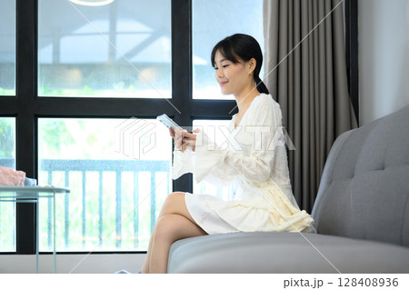 Smiling young woman in a white dress sitting on a sofa and using a smartphone 128408936