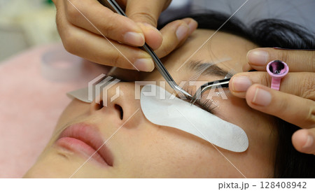 Close up of beauty salon technician applying individual eyelash extensions with precision tweezers 128408942