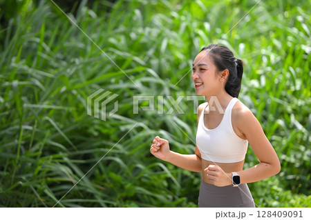Fit Asian woman jogging among lush greenery under bright natural light Fit Asian woman jogging among lush greenery under bright natural light 128409091