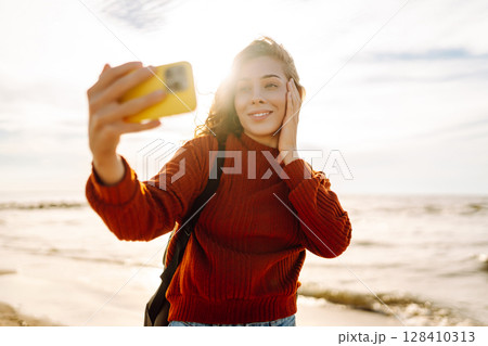 A joyful young woman takes a selfie on a sunny beach at sunset while enjoying the ocean waves. 128410313