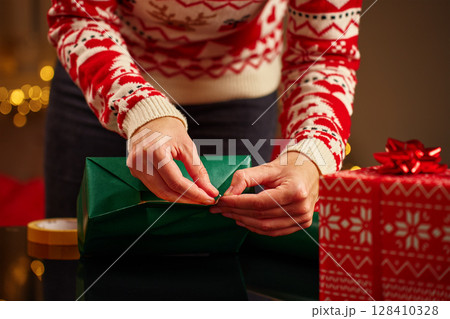 Female hands wrapping Christmas gifts in red and green paper. Woman in festive sweater preparing presents for winter holidays. Christmas and Happy New Year family traditions 128410328