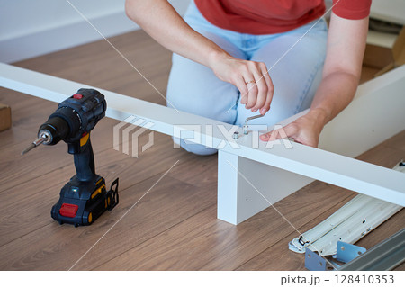 Woman assembling bed frame on wooden floor, using wrench and drill for installation. DIY furniture assembly and home improvement 128410353