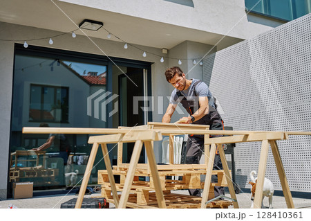 Man working on outdoor carpentry task, measuring wooden board on sawhorses in backyard Man working on outdoor carpentry task, measuring wooden board on sawhorses in backyard 128410361