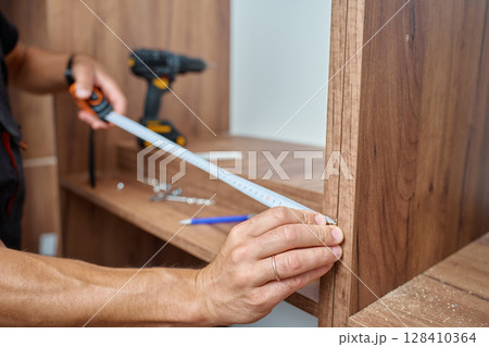 Close-up of man measuring wooden wardrobe with tape measure during furniture assembly, with tools and screws on shelf in background. Concept of home renovation and DIY furniture project 128410364