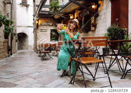 Young woman holding a smartphone in her hands and taking a selfie while sitting at the summer cafe. Young woman holding a smartphone in her hands and taking a selfie while sitting at the summer cafe. 128410729
