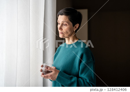 Exhausted woman with water medicine by window in stillness alone. Emotional tress, quiet reflection Exhausted woman with water medicine by window in stillness alone. Emotional tress, quiet reflection 128412406