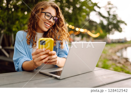 Cheerful woman sitting at a table by the lake with a laptop. Freelancing, technology concept. 128412542