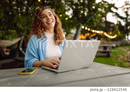 Cheerful woman in casual clothes sitting at a table by lake with laptop. Freelancing concept. Cheerful woman in casual clothes sitting at a table by lake with laptop. Freelancing concept. 128412548