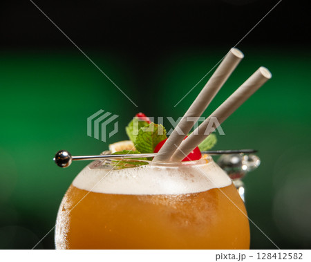 ibrant orange alcoholic cocktail, served in a unique round glass with a stem, is garnished with mint, berries, and two straws, all set on a bar counter 128412582