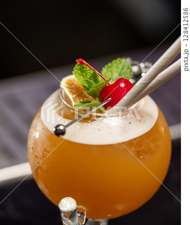 ibrant orange alcoholic cocktail, served in a unique round glass with a stem, is garnished with mint, berries, and two straws, all set on a bar counter 128412586