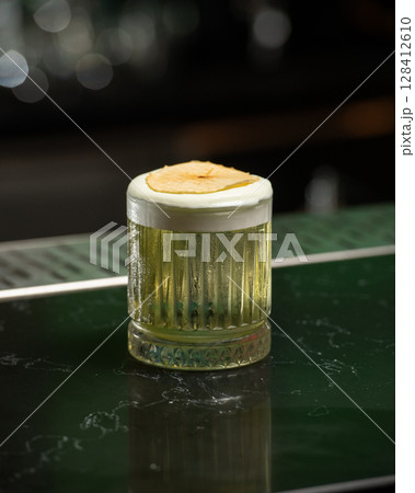 refreshing yellow-green alcoholic cocktail, topped with a white foam and a dehydrated apple slice, is served in a textured glass on a dark green bar counte 128412610