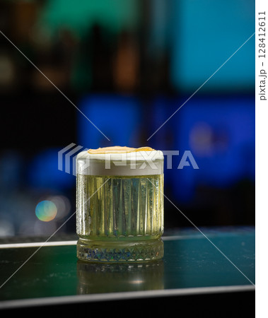 refreshing yellow-green alcoholic cocktail, topped with a white foam and a dehydrated apple slice, is served in a textured glass on a dark green bar counte 128412611
