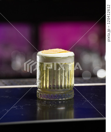 refreshing yellow-green alcoholic cocktail, topped with a white foam and a dehydrated apple slice, is served in a textured glass on a dark green bar counte 128412612