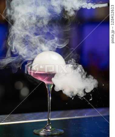 This visually striking pink alcoholic cocktail in a coupe glass features a large, dome-shaped smoke bubble on top, with more smoke billowing around it 128412613