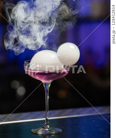 This visually striking pink alcoholic cocktail in a coupe glass features a large, dome-shaped smoke bubble on top, with more smoke billowing around it 128412614