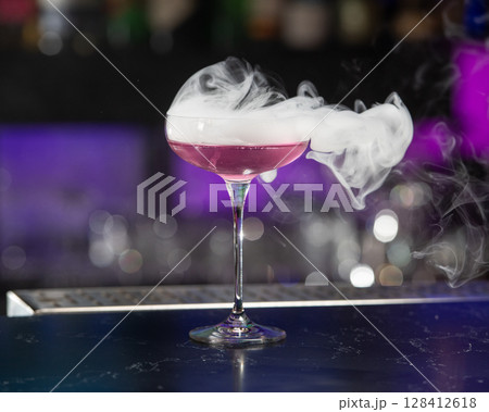 This visually striking pink alcoholic cocktail in a coupe glass features a large with more smoke billowing around it 128412618