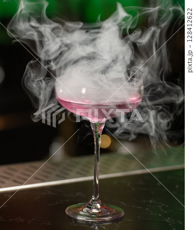 This visually striking pink alcoholic cocktail in a coupe glass features a large, dome-shaped smoke bubble on top, with more smoke billowing around it 128412622