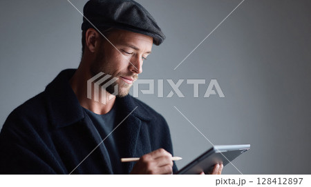 Stylish man in newsboy cap thoughtfully uses digital tablet, showcasing modern professionalism and creative focus.  Perfect for concepts of design, technology, or business. 128412897