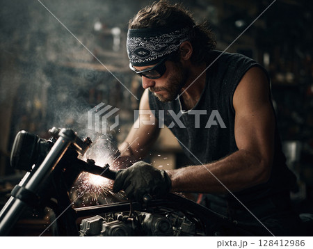 Focused mechanic welds a motorcycle frame in a dimly lit garage. Sparking action shot. Represents craftsmanship, skill, and the open road lifestyle. Grit  determination. Focused mechanic welds a motorcycle frame in a dimly lit garage. Sparking action shot. Represents craftsmanship, skill, and the open road lifestyle. Grit  determination. 128412986