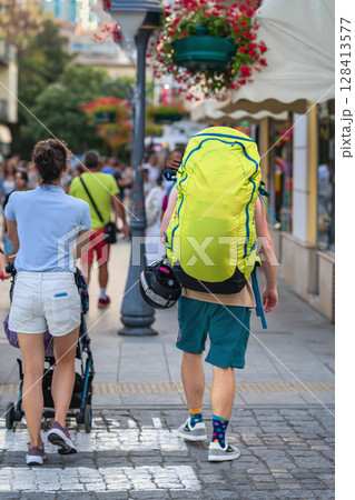 Man with oversized hiking backpack walking through lively pedestrian street in summer. Concept of adventure, travel and active lifestyle Man with oversized hiking backpack walking through lively pedestrian street in summer. Concept of adventure, travel and active lifestyle 128413577