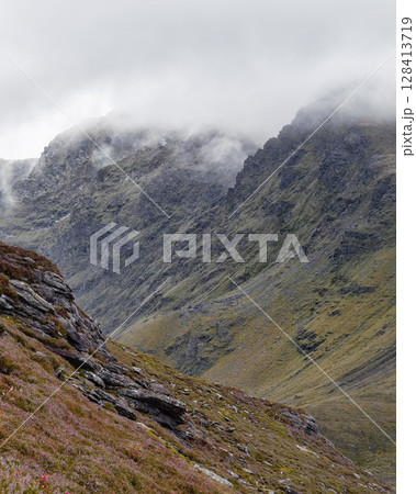 Mountains disappear into the mist, showing the rocky terrain and grassy slopes. The landscape features some heather on a cloudy day near Coomloughra Horseshoe in Ireland. 128413719