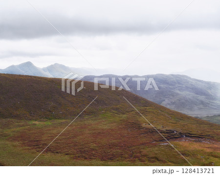 Hills are partially covered in brown and green vegetation. Mountains are visible in the distance beneath a cloudy and overcast sky in this Irish landscape. Hills are partially covered in brown and green vegetation. Mountains are visible in the distance beneath a cloudy and overcast sky in this Irish landscape. 128413721