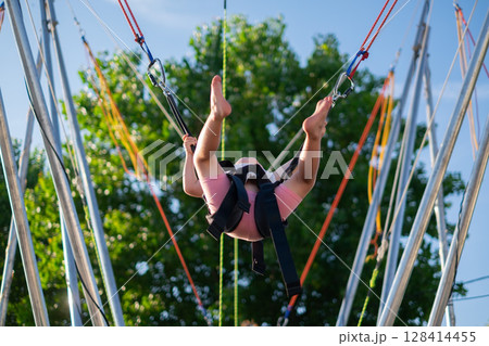 Little European Girl Learning to Jump on a Trampoline with Safety Harness. Summer Fun Little European Girl Learning to Jump on a Trampoline with Safety Harness. Summer Fun 128414455