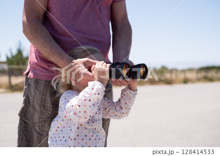 A girl looks into the distance through binoculars. Her grandfather helps her A girl looks into the distance through binoculars. Her grandfather helps her 128414533