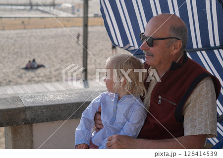 An elderly Spanish man sits with his granddaughter under a parasol on a promenade  128414539