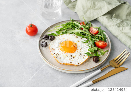 Fried eggs with arugula, ovives and cherry tomatoes on a plate on a light background Fried eggs with arugula, ovives and cherry tomatoes on a plate on a light background 128414984