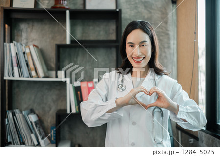 A cheerful doctor in a white coat making a heart gesture, standing in a modern office with a bookshelf and natural light. A cheerful doctor in a white coat making a heart gesture, standing in a modern office with a bookshelf and natural light. 128415025