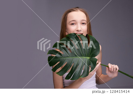 Young caucasian female teen with long hair holding large monstera leaf against Young caucasian female teen with long hair holding large monstera leaf against 128415414