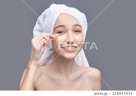 Young caucasian female applying under eye patch with towel on head against gray Young caucasian female applying under eye patch with towel on head against gray 128415432