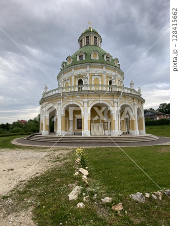 Baroque-style church with green dome framed by cloudy sky and vibrant greenery 128415662