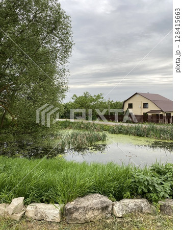 Idyllic pond with lush greenery and quaint house, offering serene natural scenery as background 128415663