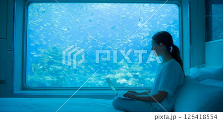 Woman working on laptop in underwater accommodation, surrounded by vibrant marine life, showcasing a unique workcation experience with stunning views 128418554