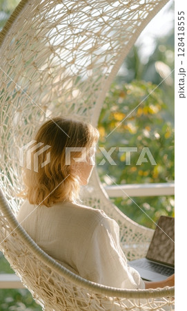 Woman working on laptop while relaxing in a hanging chair surrounded by greenery, embodying the essence of a workcation lifestyle 128418555