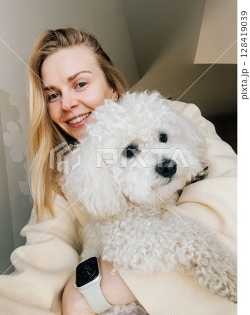 Happy young woman wearing beige sweatshirt, smiling while holding her fluffy white dog, enjoying cozy moment at home filled with warmth and affection. Cheerful blonde woman embracing fluffy white dog Happy young woman wearing beige sweatshirt, smiling while holding her fluffy white dog, enjoying cozy moment at home filled with warmth and affection. Cheerful blonde woman embracing fluffy white dog 128419039